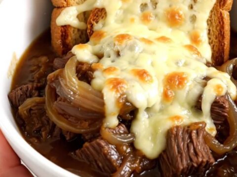 A steaming bowl of French onion beef stew with tender beef, potatoes, and onions, topped with cheesy toast.