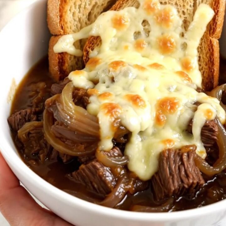 A steaming bowl of French onion beef stew with tender beef, potatoes, and onions, topped with cheesy toast.
