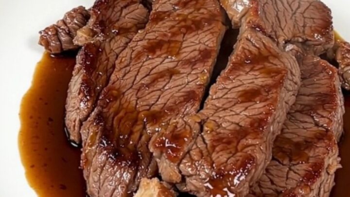 A glossy and tender ginger soy flank steak served on a white plate with a blurred kitchen background.