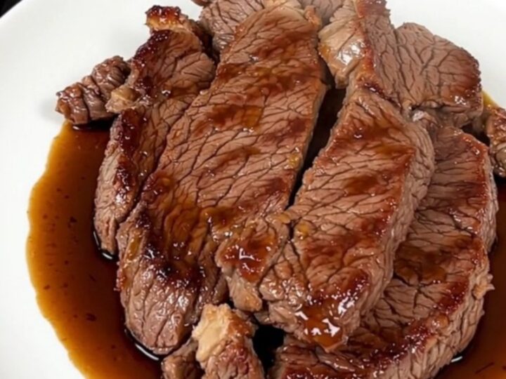 A glossy and tender ginger soy flank steak served on a white plate with a blurred kitchen background.