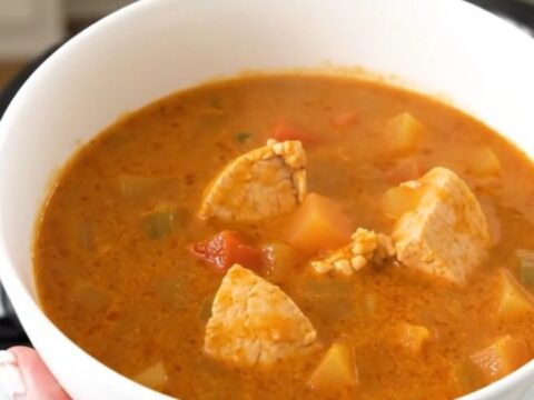 A white bowl filled with creamy tomato chicken stew, garnished with herbs, held in front of a bright kitchen background.