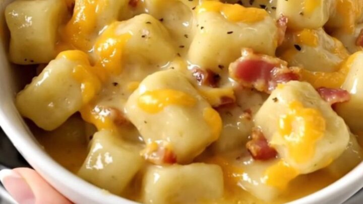 A close-up of creamy cheesy bacon gnocchi in a white bowl with a soft-focus kitchen background.