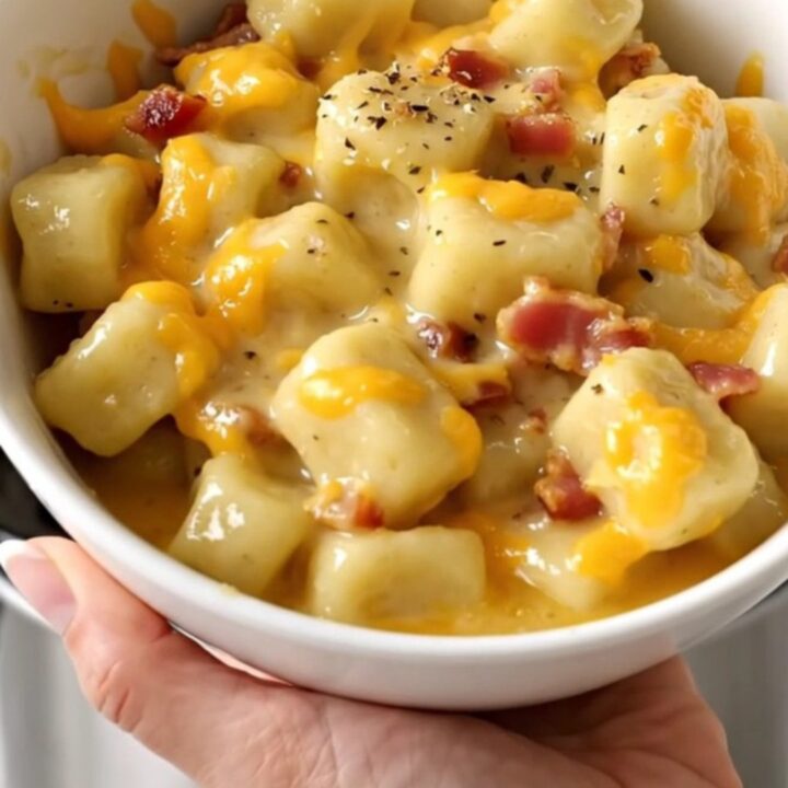 A close-up of creamy cheesy bacon gnocchi in a white bowl with a soft-focus kitchen background.