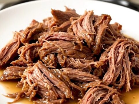 A succulent beef chuck roast sitting in a dark slow cooker, surrounded by aromatic garlic and herbs.
