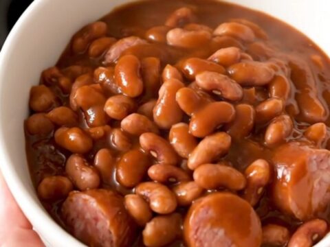 A top-down view of a black slow cooker filled with sliced kielbasa and mixed beans covered in barbecue sauce.