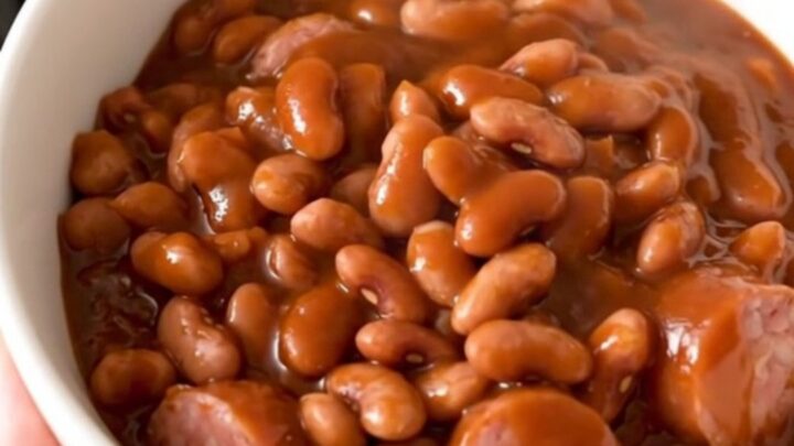 A top-down view of a black slow cooker filled with sliced kielbasa and mixed beans covered in barbecue sauce.
