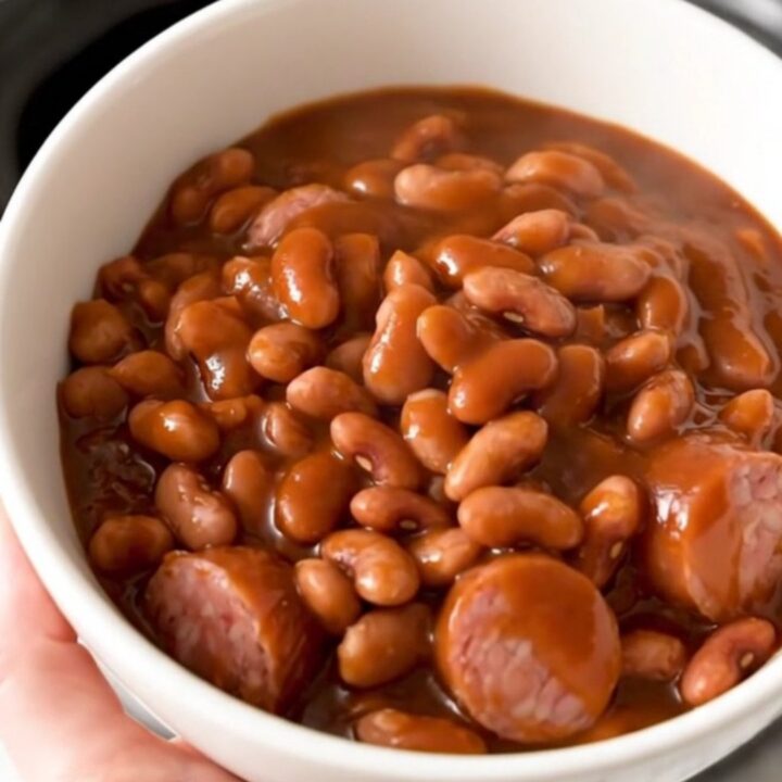 A top-down view of a black slow cooker filled with sliced kielbasa and mixed beans covered in barbecue sauce.