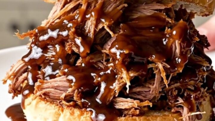 A close-up shot of glossy, steaming shredded pulled pork on a toasted bun on a plain white plate.