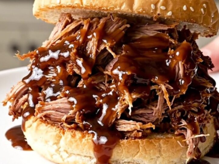 A close-up shot of glossy, steaming shredded pulled pork on a toasted bun on a plain white plate.