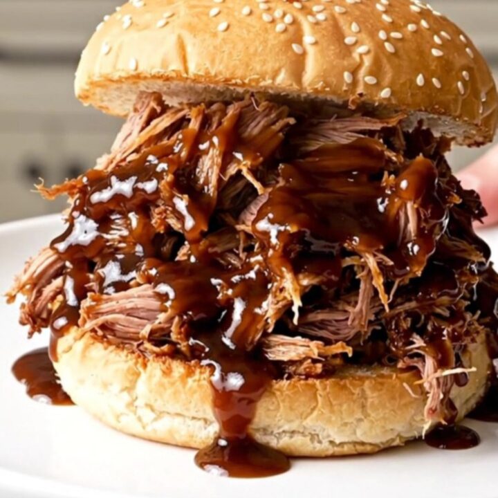 A close-up shot of glossy, steaming shredded pulled pork on a toasted bun on a plain white plate.