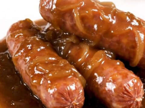 A close-up of glossy, cooked bratwurst links smothered in a rich, dark onion gravy on a white plate.