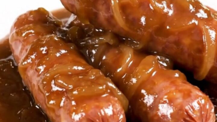 A close-up of glossy, cooked bratwurst links smothered in a rich, dark onion gravy on a white plate.