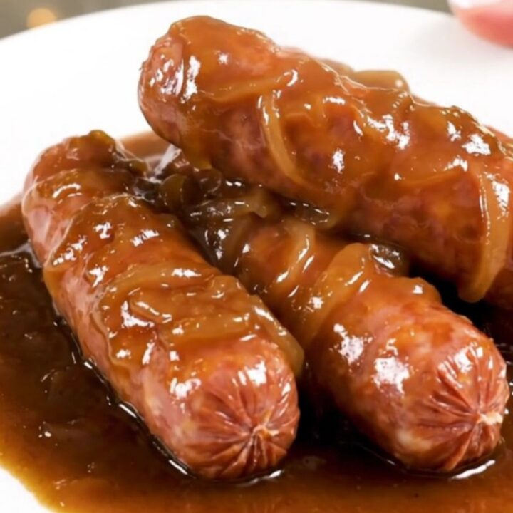 A close-up of glossy, cooked bratwurst links smothered in a rich, dark onion gravy on a white plate.