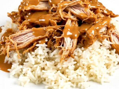 A close-up of glossy shredded Thai peanut chicken served over a bed of steaming white rice on a round plate.