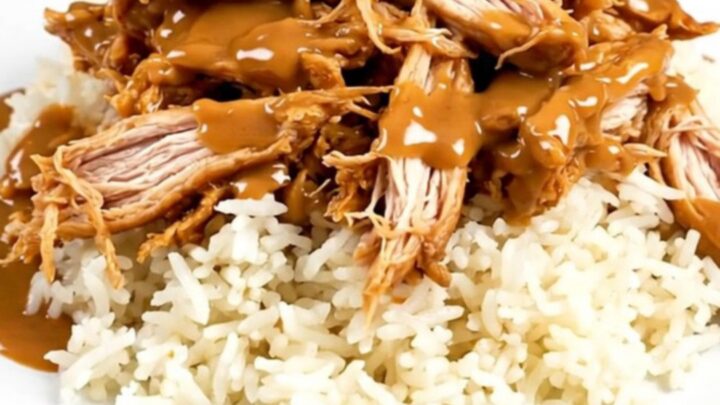 A close-up of glossy shredded Thai peanut chicken served over a bed of steaming white rice on a round plate.