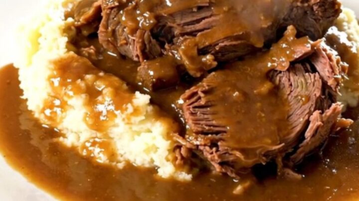 A close-up of a tender beef chuck roast in a black slow cooker with baby carrots and a rich brown mushroom gravy.