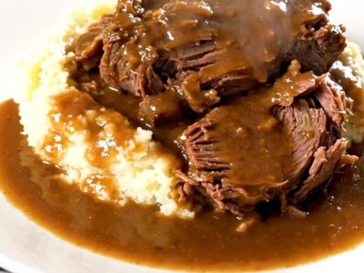 A close-up of a tender beef chuck roast in a black slow cooker with baby carrots and a rich brown mushroom gravy.