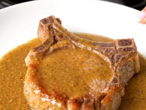 A close-up of a tender, cooked bone-in pork chop covered in a glossy, creamy Dijon and ranch sauce on a white plate.