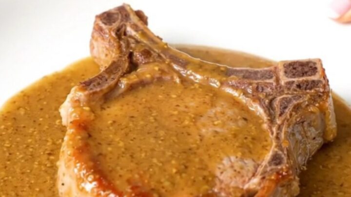 A close-up of a tender, cooked bone-in pork chop covered in a glossy, creamy Dijon and ranch sauce on a white plate.