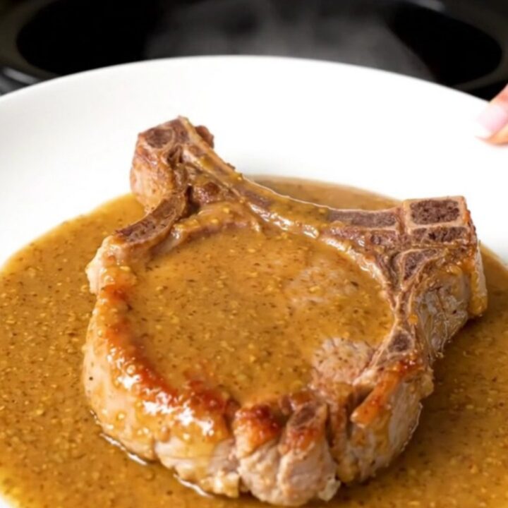 A close-up of a tender, cooked bone-in pork chop covered in a glossy, creamy Dijon and ranch sauce on a white plate.