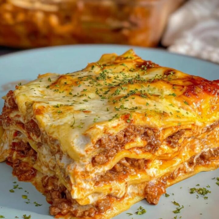 A tall, pristine square slice of creamy Alfredo beef lasagna served on a light blue plate with golden-baked cheese.