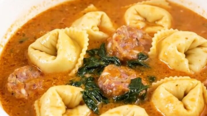 A white bowl filled with creamy spinach tortellini tomato soup, featuring visible sausage and green spinach leaves.
