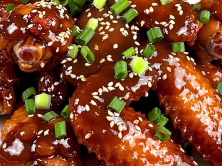 A close-up of glossy, glazed General Tso chicken wings on a white plate, garnished with scallions and sesame seeds.
