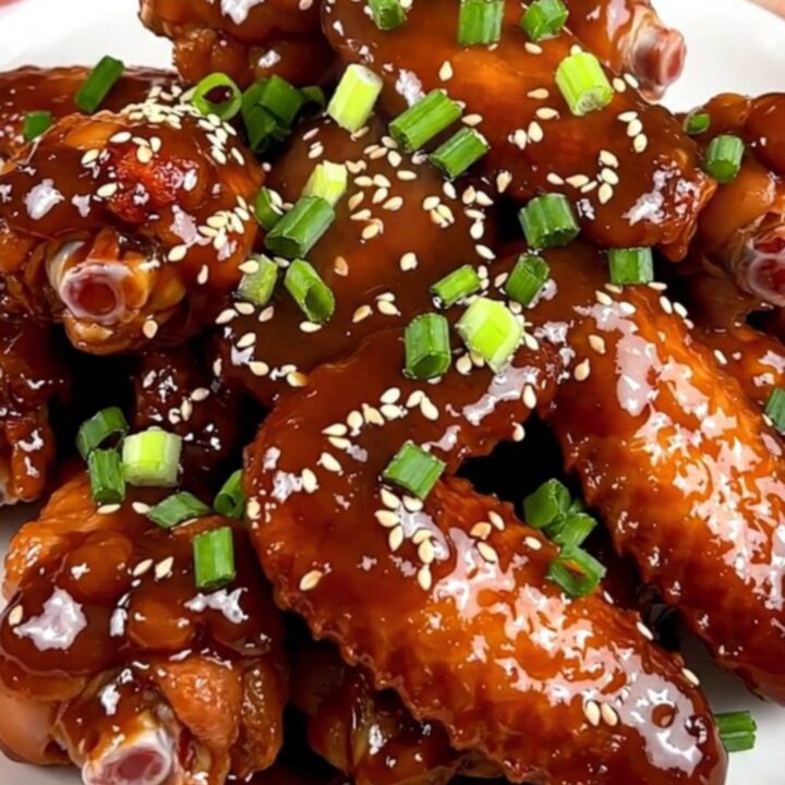 A close-up of glossy, glazed General Tso chicken wings on a white plate, garnished with scallions and sesame seeds.