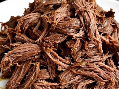 A close-up view of tender, glossy, shredded beef served on a plain white plate in a bright kitchen.