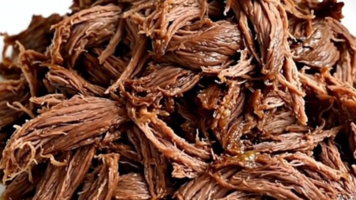 A close-up view of tender, glossy, shredded beef served on a plain white plate in a bright kitchen.