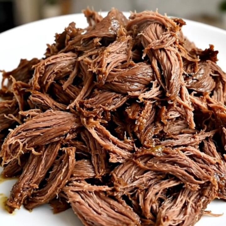 A close-up view of tender, glossy, shredded beef served on a plain white plate in a bright kitchen.