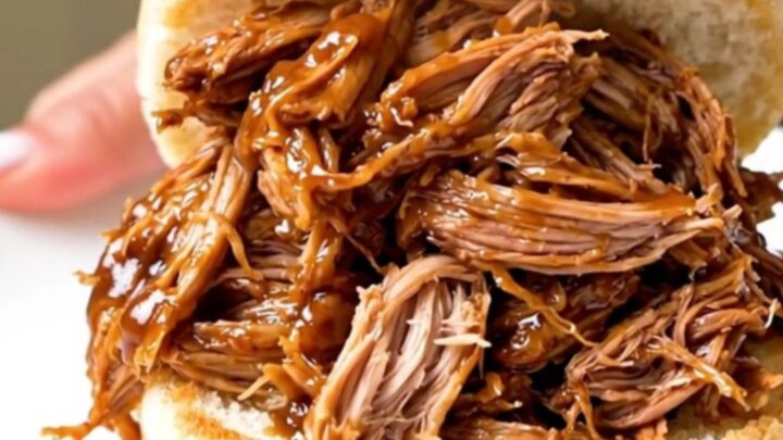 A close-up of a generous portion of shredded pulled pork piled high on a soft bun, showing glossy sauce and tender meat.