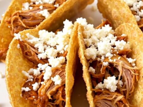 A close-up of shredded honey lime chicken in a crispy taco shell topped with crumbled white cotija cheese.