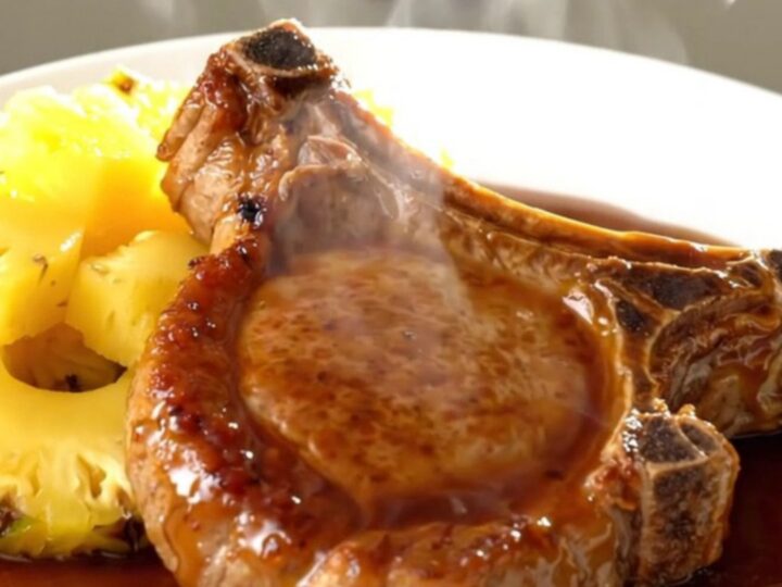 A close-up of a tender, glossy teriyaki glazed pork chop with pineapple chunks on a white plate.