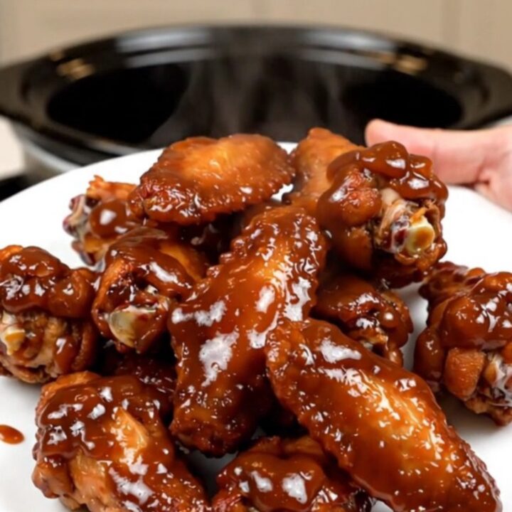 A top-down view of raw chicken wings and onions being prepared in a black slow cooker on a white countertop.