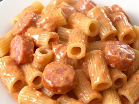 A close-up of a white plate filled with creamy Italian sausage and rigatoni pasta, steaming and topped with fresh herbs.