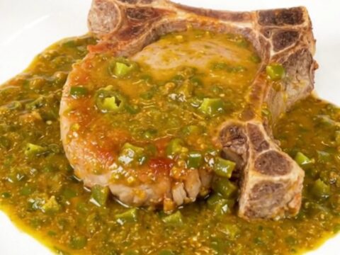 A close-up view of a tender pork chop covered in rich, creamy green chile queso sauce on a clean white plate.