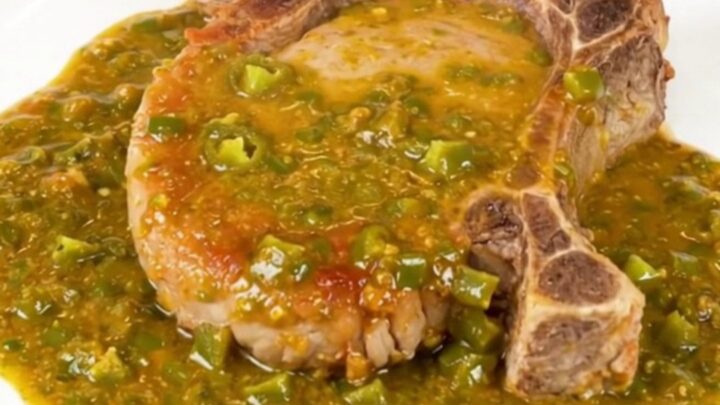A close-up view of a tender pork chop covered in rich, creamy green chile queso sauce on a clean white plate.