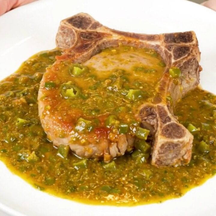 A close-up view of a tender pork chop covered in rich, creamy green chile queso sauce on a clean white plate.
