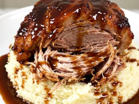 A close-up of a tender, shredded honey balsamic pork roast served over a portion of creamy mashed potatoes on a white plate.