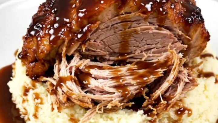 A close-up of a tender, shredded honey balsamic pork roast served over a portion of creamy mashed potatoes on a white plate.