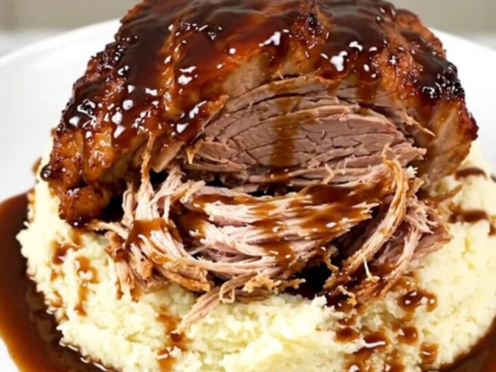A close-up of a tender, shredded honey balsamic pork roast served over a portion of creamy mashed potatoes on a white plate.
