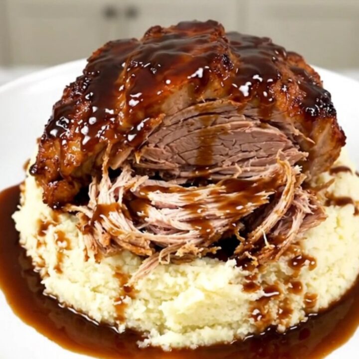 A close-up of a tender, shredded honey balsamic pork roast served over a portion of creamy mashed potatoes on a white plate.
