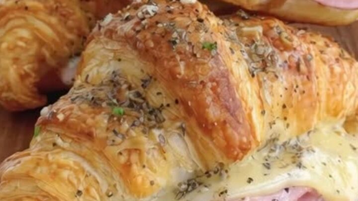 Three warm, golden-brown ham and Swiss croissants stacked on a rustic wooden board in a sunlit kitchen.
