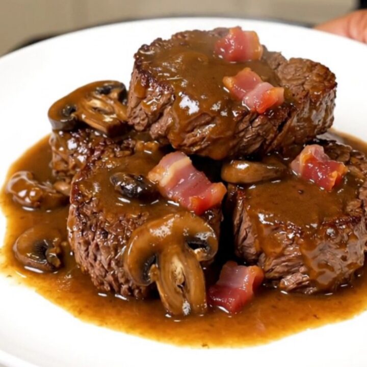A close-up of tender, glossy beef chuck cubes served on a white plate with sautéed mushrooms and bacon.