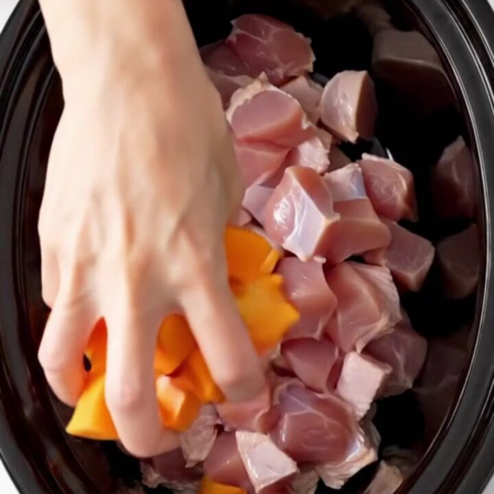 A top-down shot of a black slow cooker filled with cubed turkey, butternut squash, and cranberry sauce before cooking.