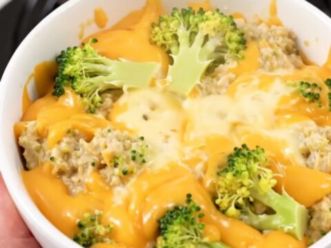 A top-down view of a black slow cooker filled with layers of fresh broccoli, shredded cheddar, and cubes of cream cheese.