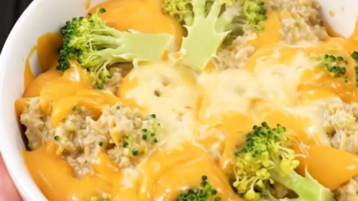 A top-down view of a black slow cooker filled with layers of fresh broccoli, shredded cheddar, and cubes of cream cheese.