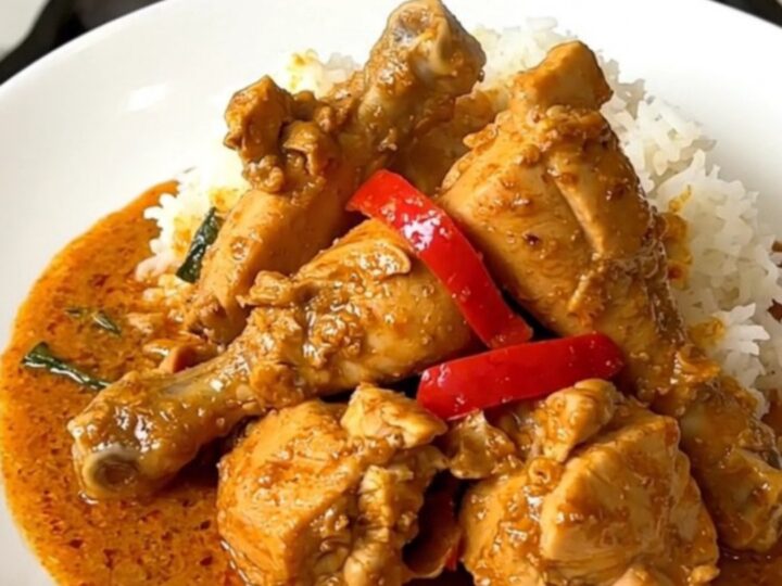 A top-down view of a black slow cooker containing raw chicken, sliced red peppers, and coconut milk.