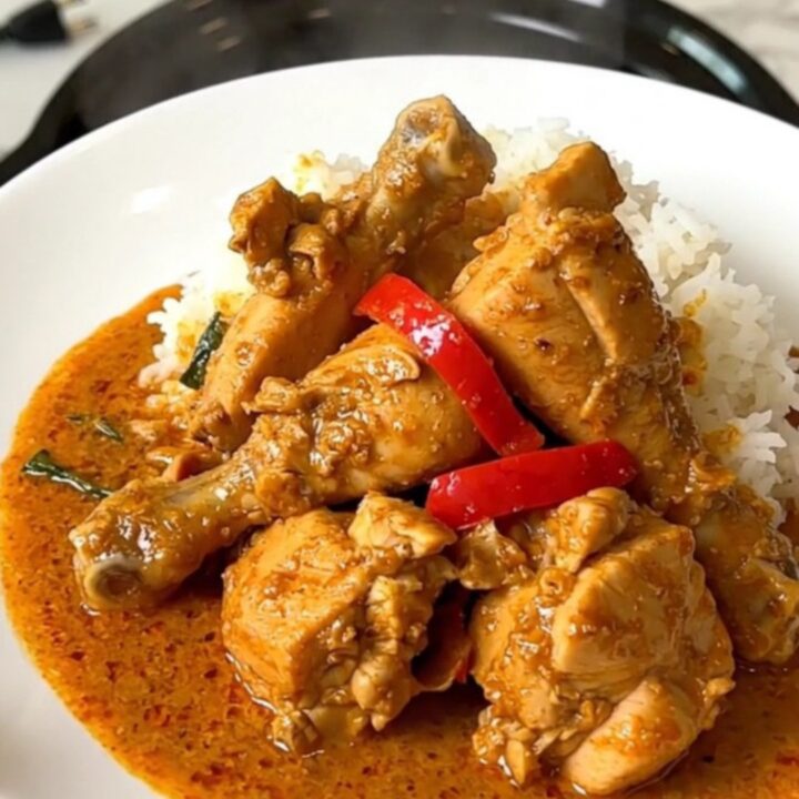 A top-down view of a black slow cooker containing raw chicken, sliced red peppers, and coconut milk.